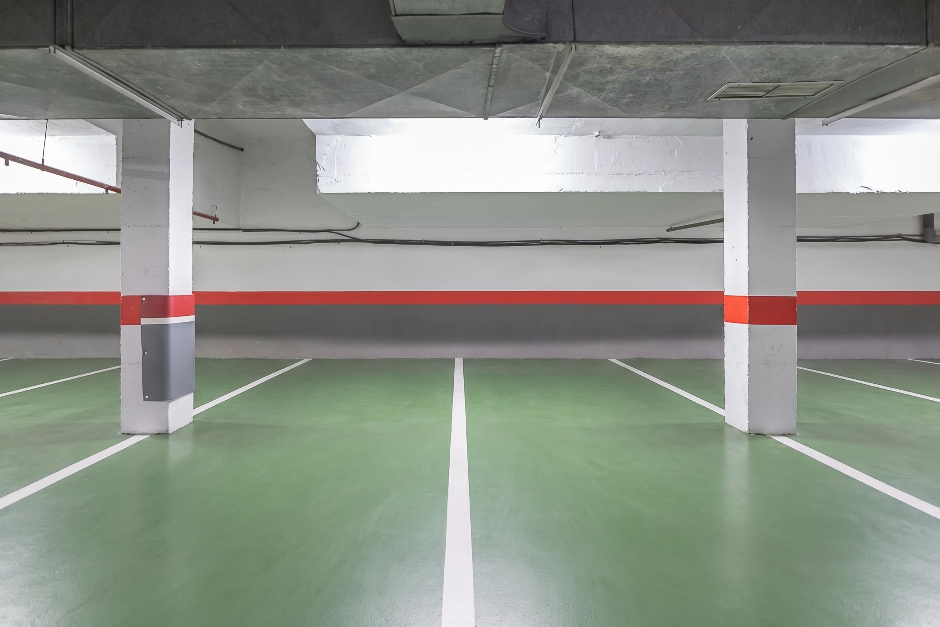 Un estacionamiento interior con piso verde, líneas pintadas de blanco y rojo, y pilares de concreto.