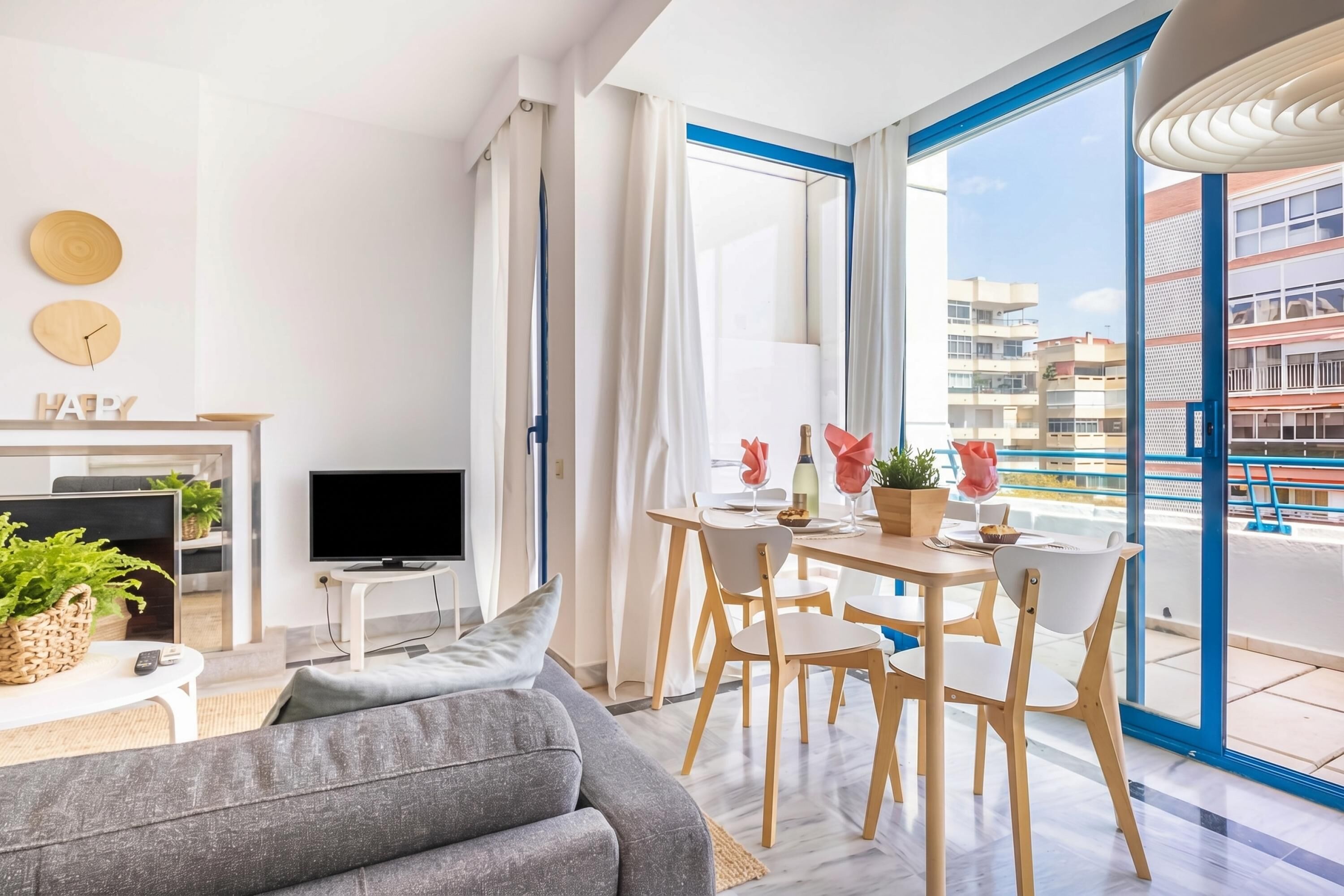 A modern living room with a sofa, TV, dining table, chairs, and a balcony with a view of buildings.