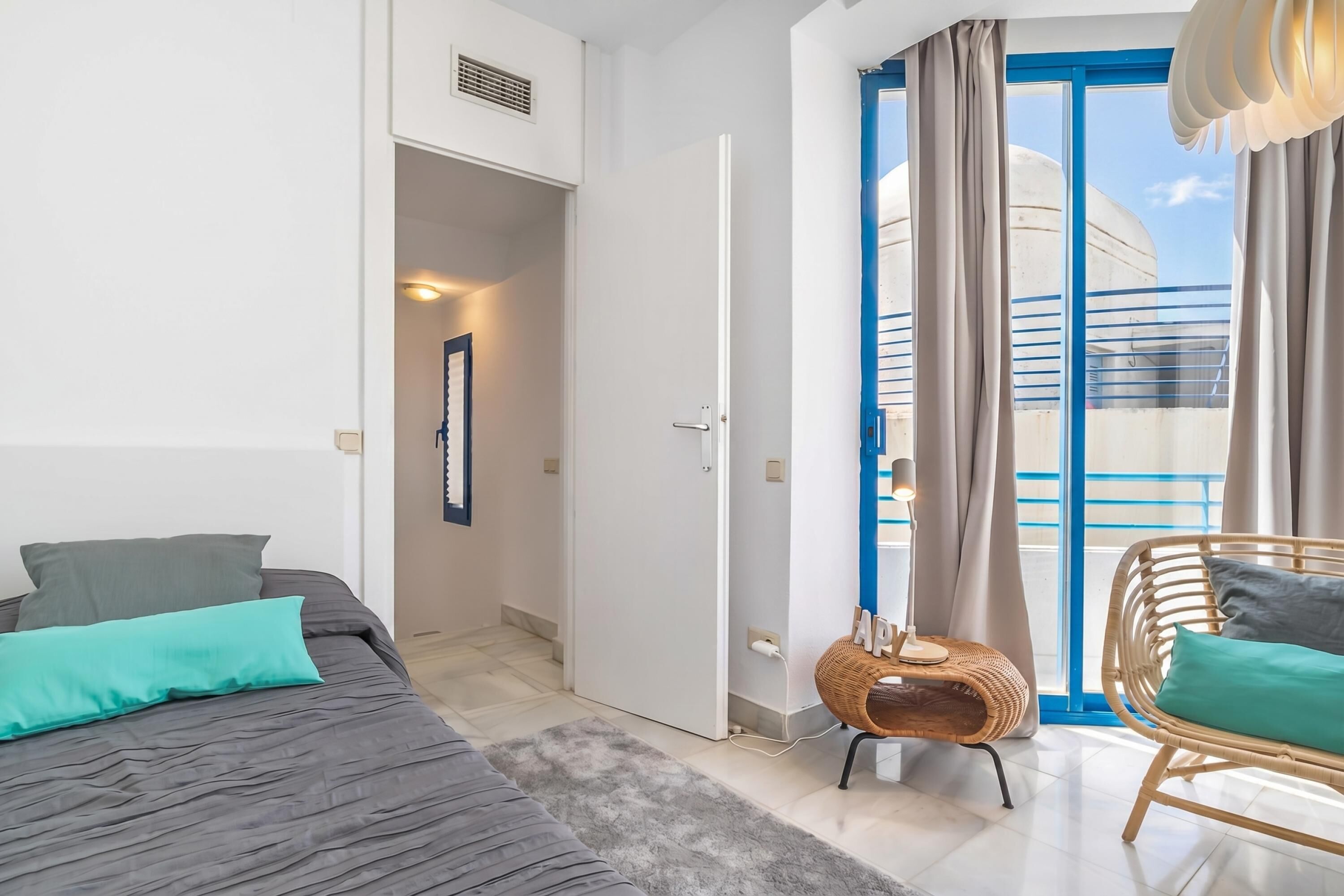 A modern hotel room with a bed, a wicker chair, a balcony with blue railings, and a view of a building.
