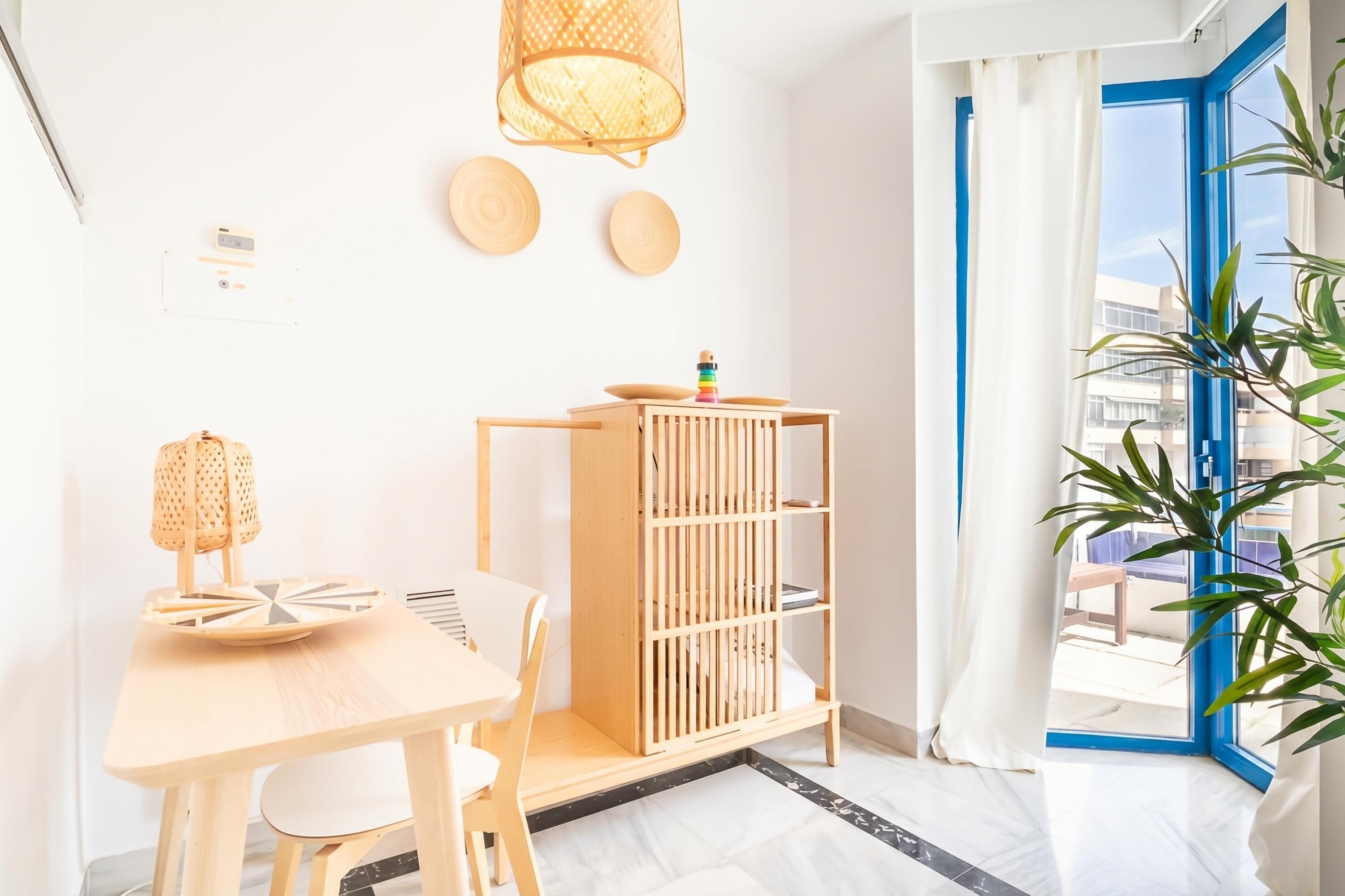 A room with a wooden table, chairs, a bookshelf, a hanging lamp, and a window with blue trim.