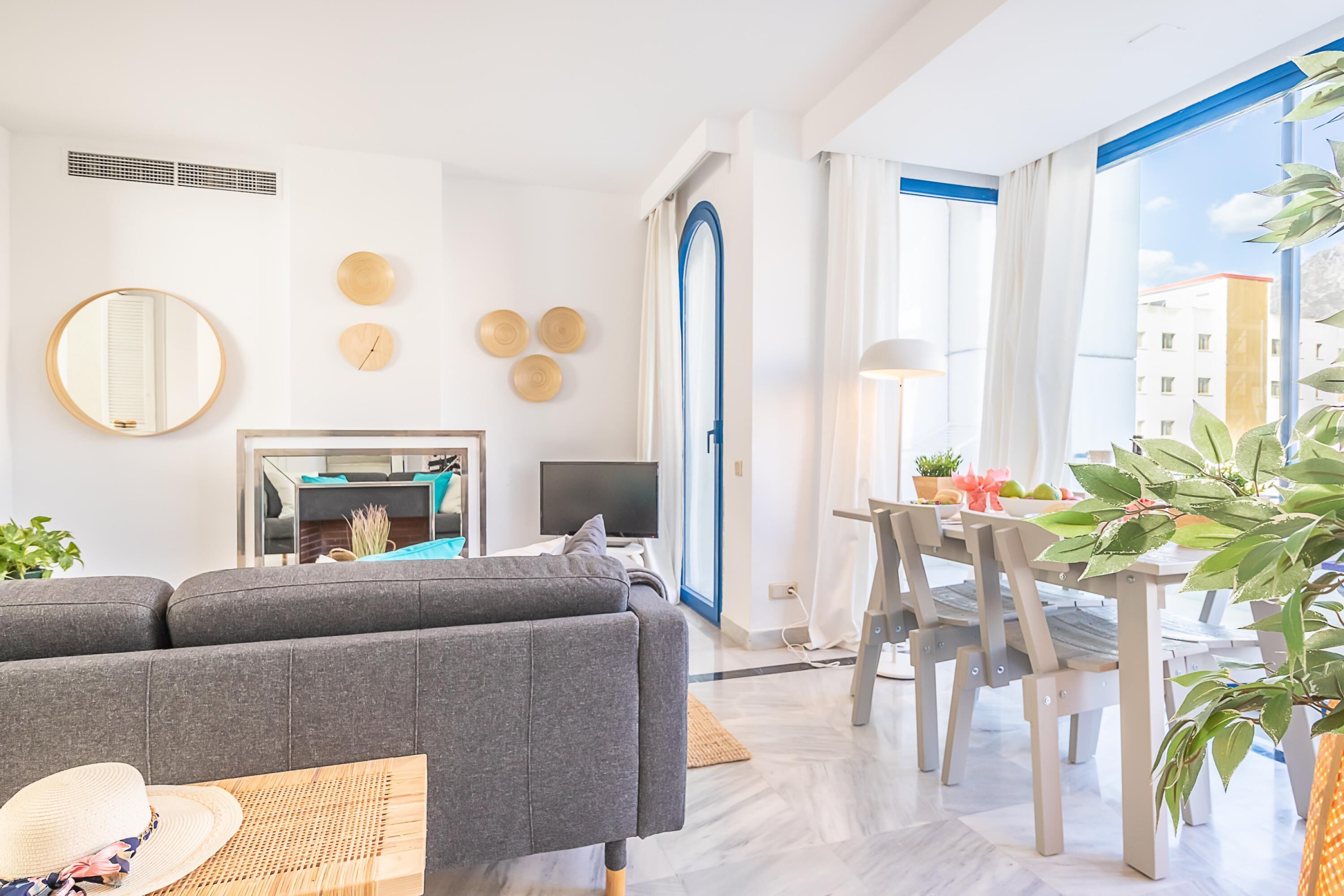 Un salon moderne avec un canapé gris, une table à manger et des chaises. On y trouve également un téléviseur, un miroir et des œuvres d’art décoratives accrochées au mur.