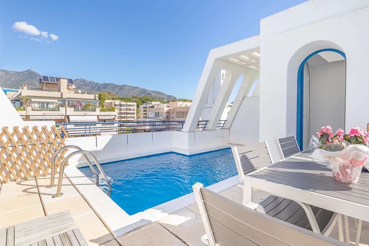 A pool area with a table and chairs, a wooden deck, and a view of buildings and mountains in the background.