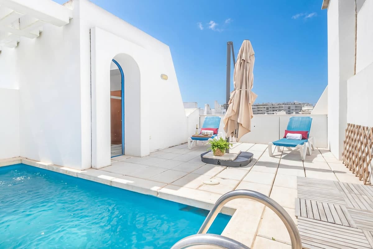 A rooftop pool area with lounge chairs, a small table with a plant, and a view of the city skyline.