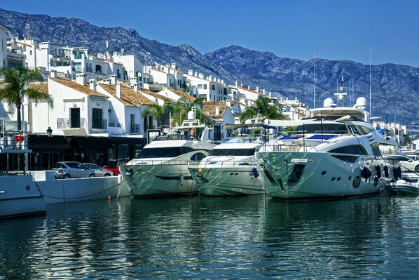 A marina with several yachts docked, white buildings with red-tiled roofs, and palm trees in the foreground.