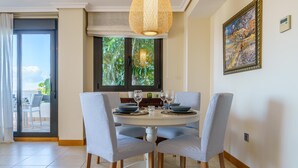 A dining area with a round table, four chairs, a hanging light fixture, a window with a view, and a painting on the wall.