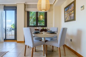 A dining area with a round table, four chairs, a hanging light fixture, a window with a view, and a painting on the wall. - Sunset Seaview Corner With Pool (Estepona)