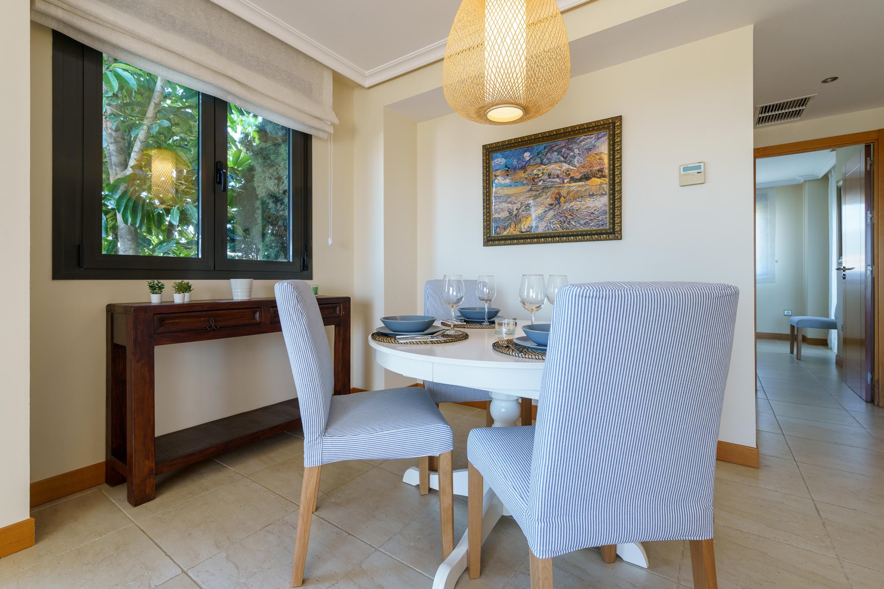 A dining area with a round table, chairs, a painting, and a window with a view of greenery.