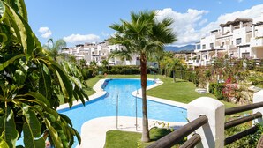 A swimming pool with a curved design, surrounded by a well-maintained lawn, palm trees, and a row of white buildings with balconies.