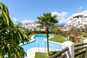 A swimming pool with a curved design, surrounded by a well-maintained lawn, palm trees, and a row of white buildings with balconies. - Sunset Seaview Corner With Pool (Estepona)