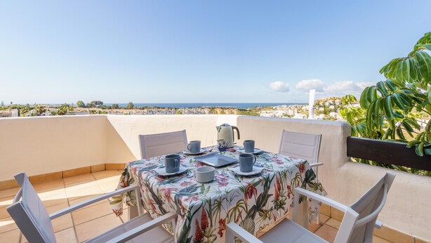 A table set for two with a floral tablecloth, a kettle, and cups on a balcony overlooking a coastal view.