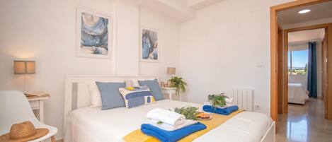 A hotel room with a bed, a chair, artwork on the walls, and a view through an open door.