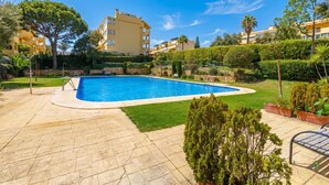 Una piscina con solárium de piedra, rodeada de césped y setos cuidados, con un edificio al fondo.