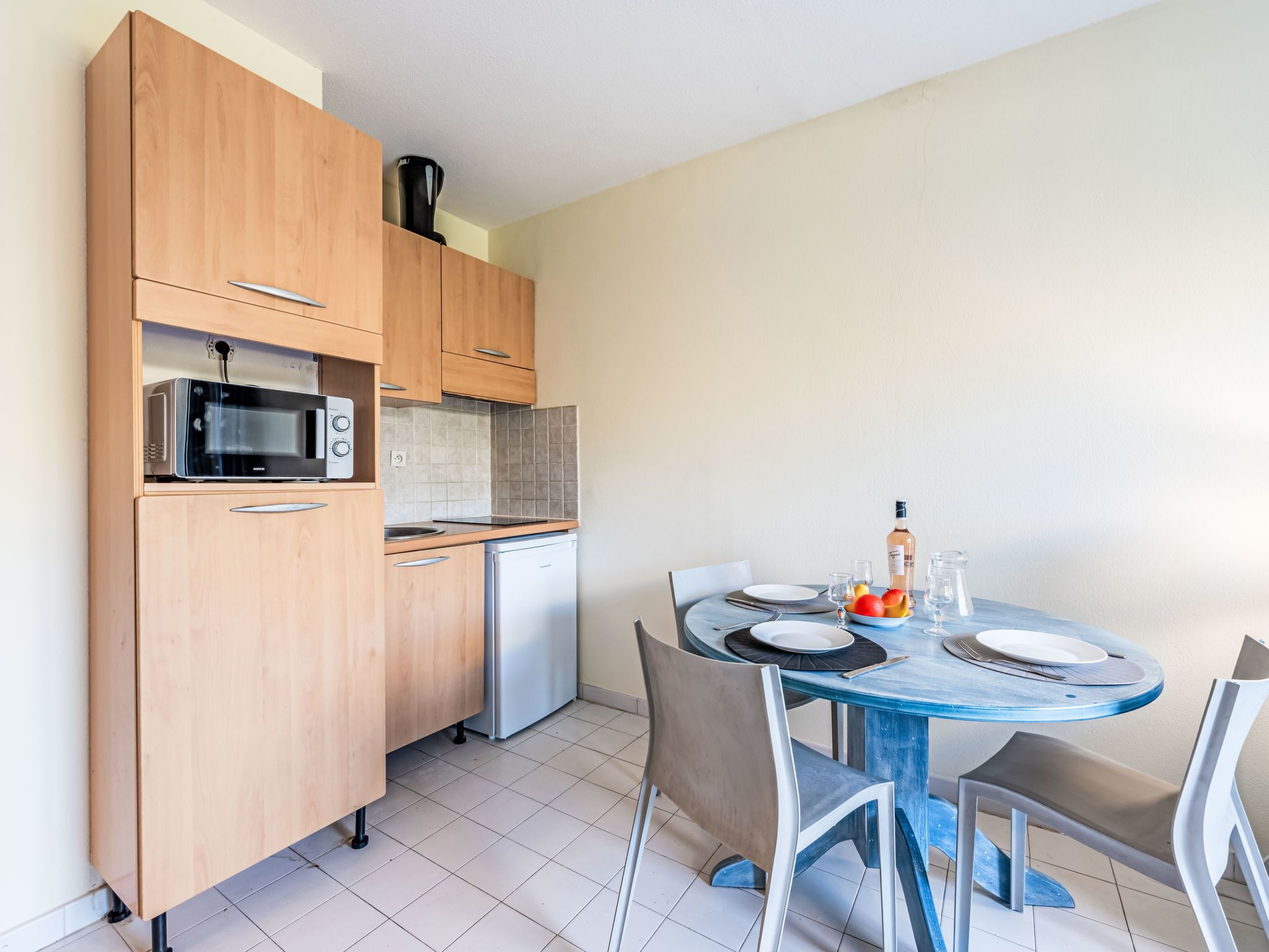 Cocina compacta con armarios de madera, microondas y refrigerador pequeño. Hay una mesa redonda con sillas y un comedor.