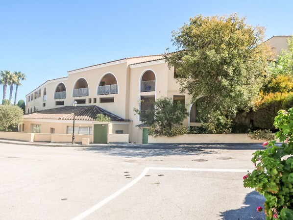 A multi-story building with arched windows and balconies, a flat roof, and a paved parking area.