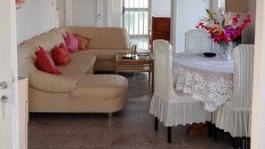 Un salon avec un canapé beige, une table à manger recouverte d’une nappe blanche en dentelle et un luminaire suspendu rouge.