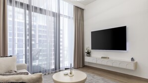 Un salon moderne doté d’un grand téléviseur à écran plat fixé au mur, d’une table basse ronde, d’un canapé et offrant une vue sur la ville grâce à de grandes fenêtres ornées de rideaux légers.