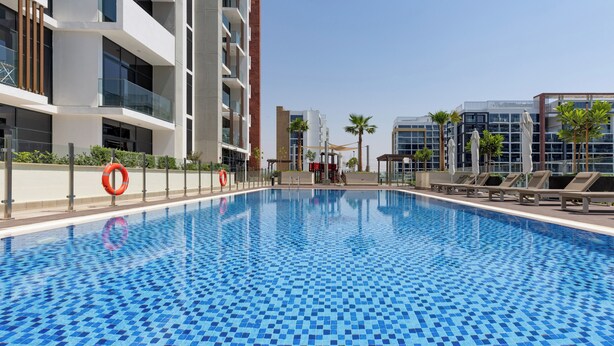Une piscine dont le fond est recouvert de carreaux disposés en motif à damier, entourée de bâtiments modernes et de palmiers.