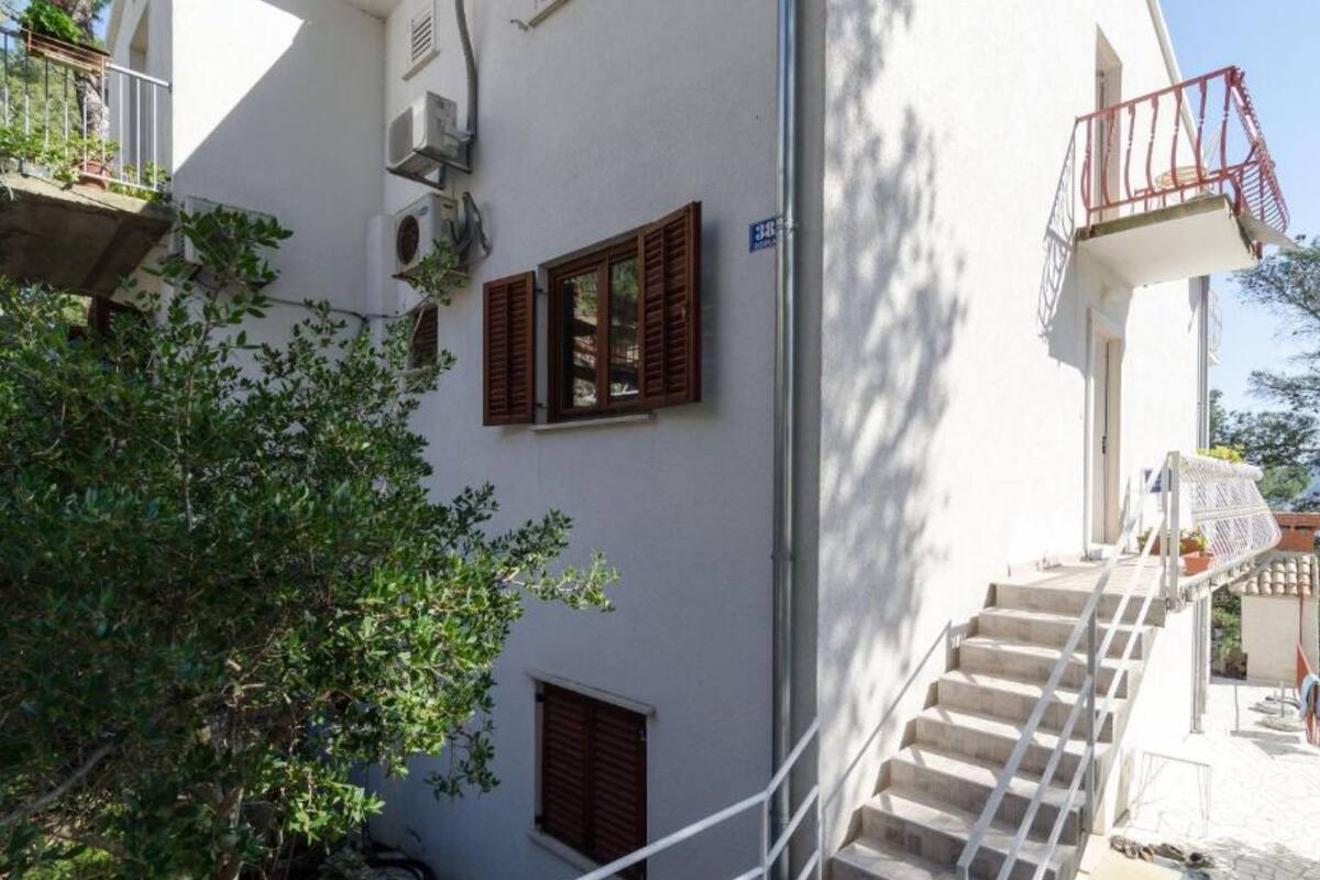 A white building with a staircase leading to a balcony. There are two windows with shutters, and an air conditioning unit is mounted on the wall.