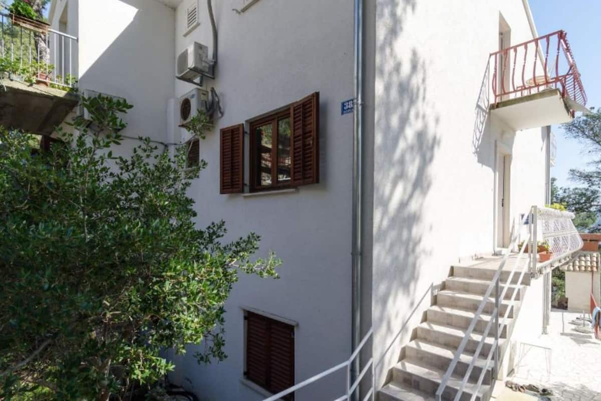 A white building with a staircase leading to a balcony. There are two windows with shutters, and an air conditioning unit is mounted on the wall.