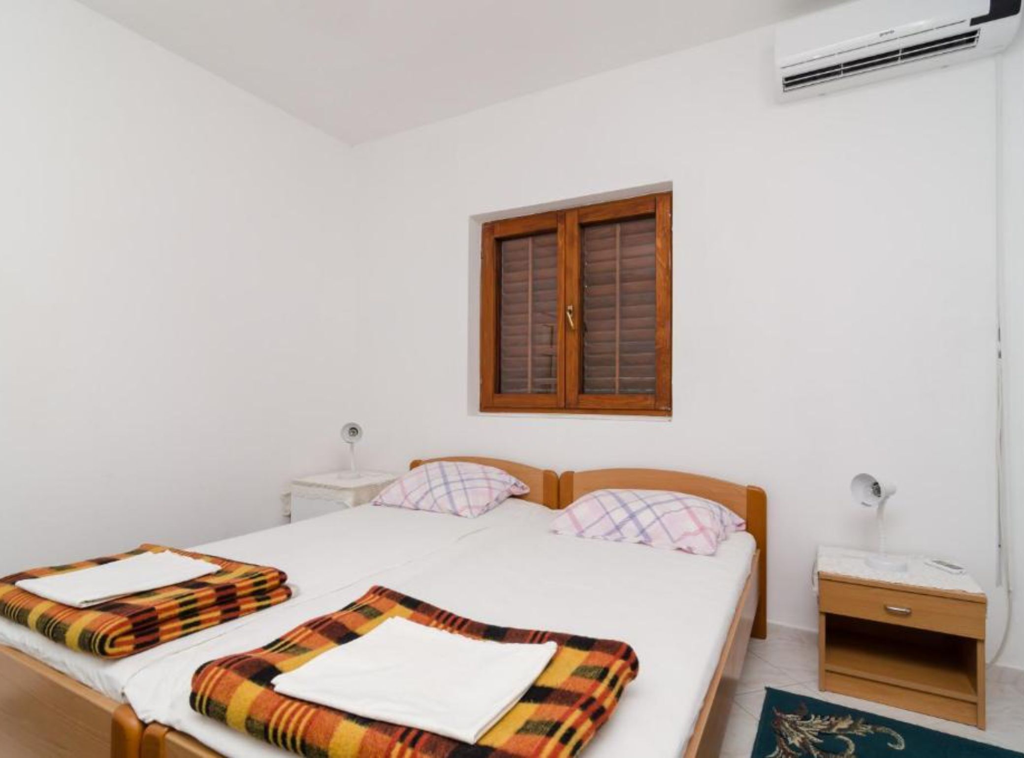 A double bed with a wooden headboard, two bedside tables, a window with wooden shutters, and an air conditioning unit.