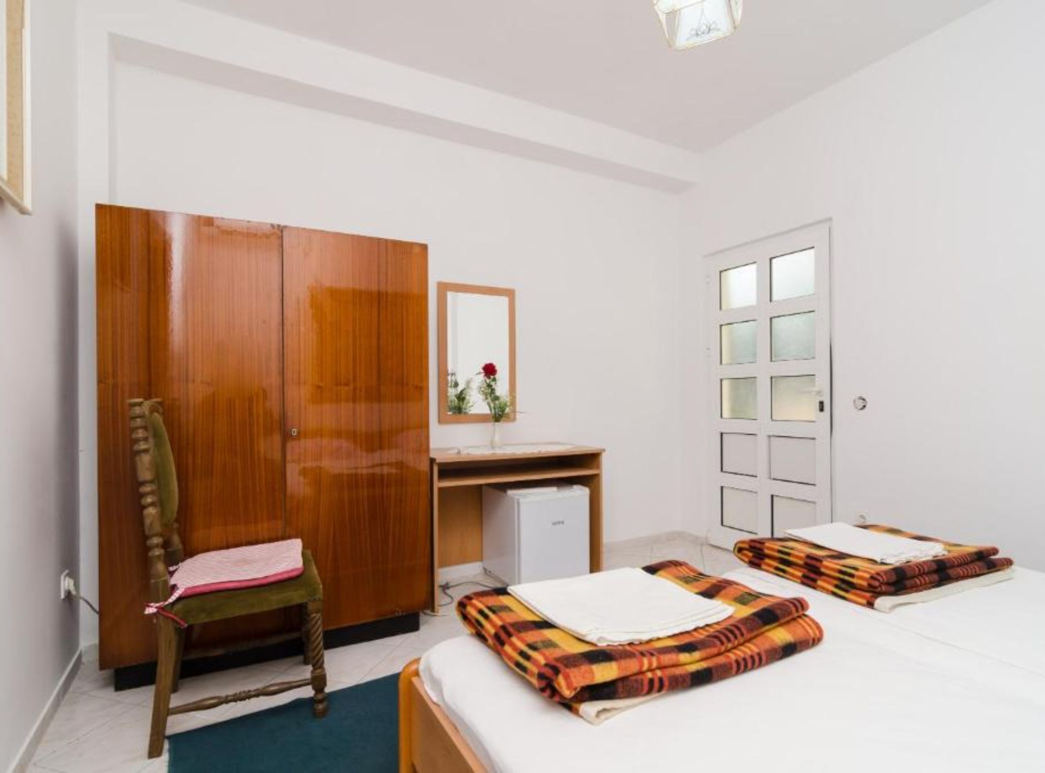 A bedroom with a wooden wardrobe, a desk with a mirror, a bed with a plaid blanket, and a small bedside table.