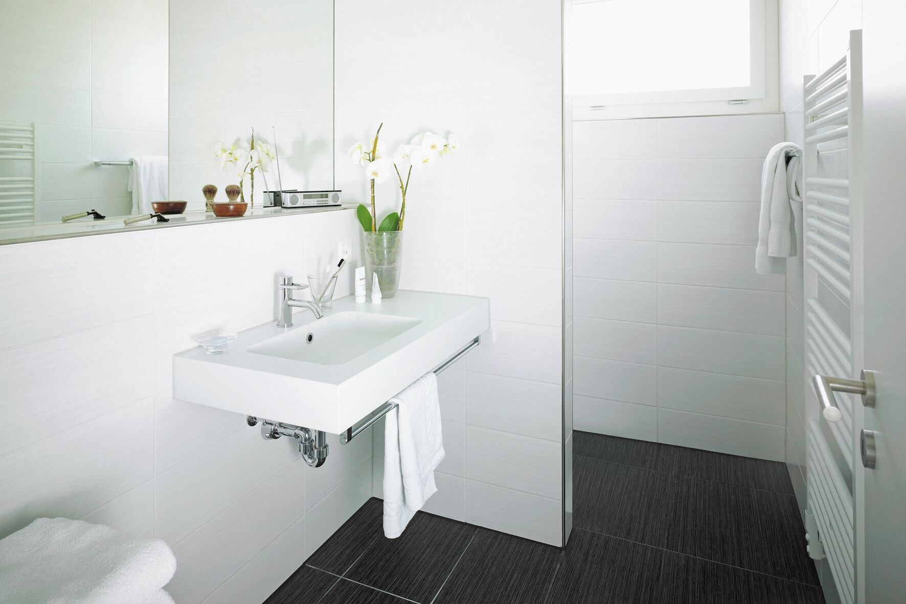 Un baño moderno con lavamanos, espejo y azulejos blancos. Toallas blancas cuelgan de la pared y una puerta de cristal para la ducha.