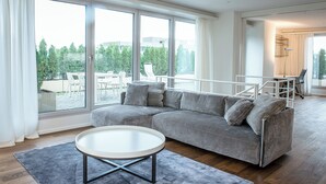 En moderne stue med en grå sofa, et rundt sofabord og store vinduer med udsigt til udendørs møbler og grønne planter.