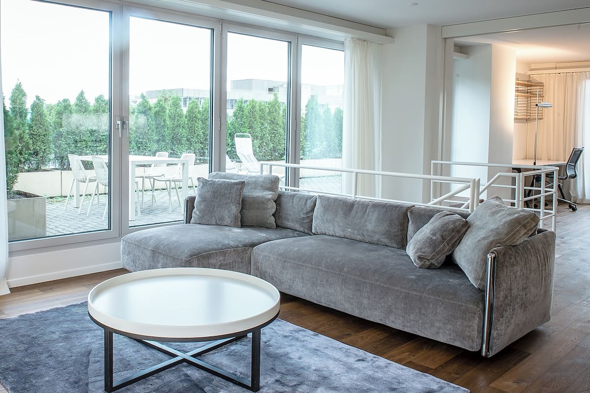 A modern living room with a grey sofa, a round coffee table, and large windows offering a view of outdoor furniture and greenery.