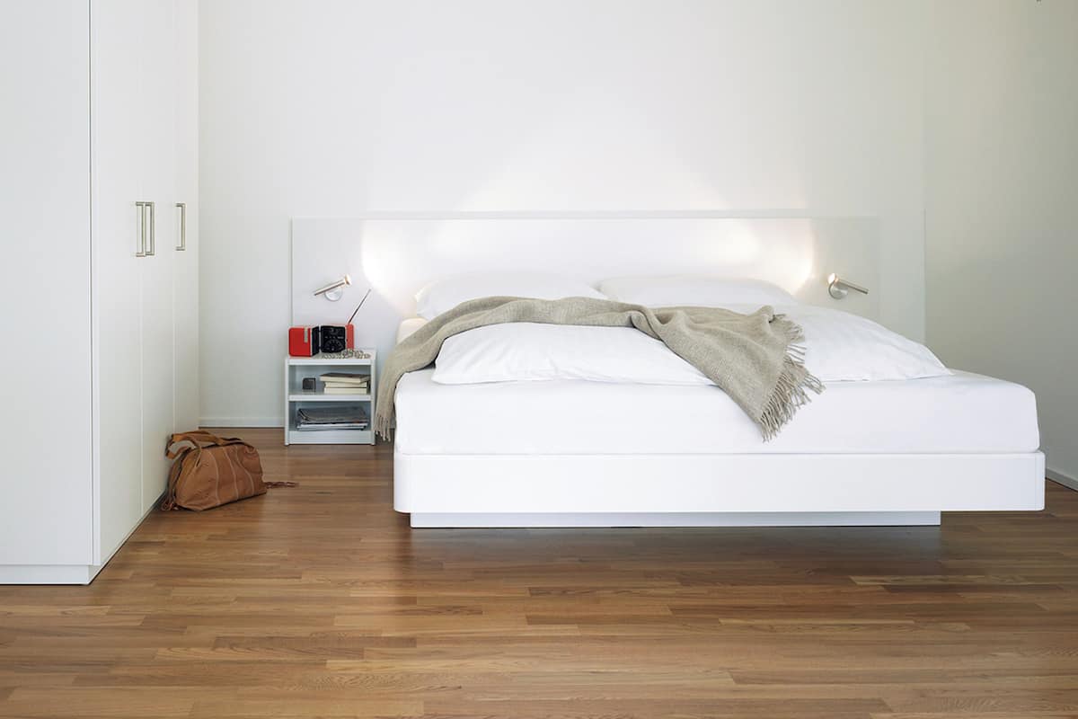 A modern bedroom with a large bed, a bedside table with a lamp, and a small shelf with books and a radio.