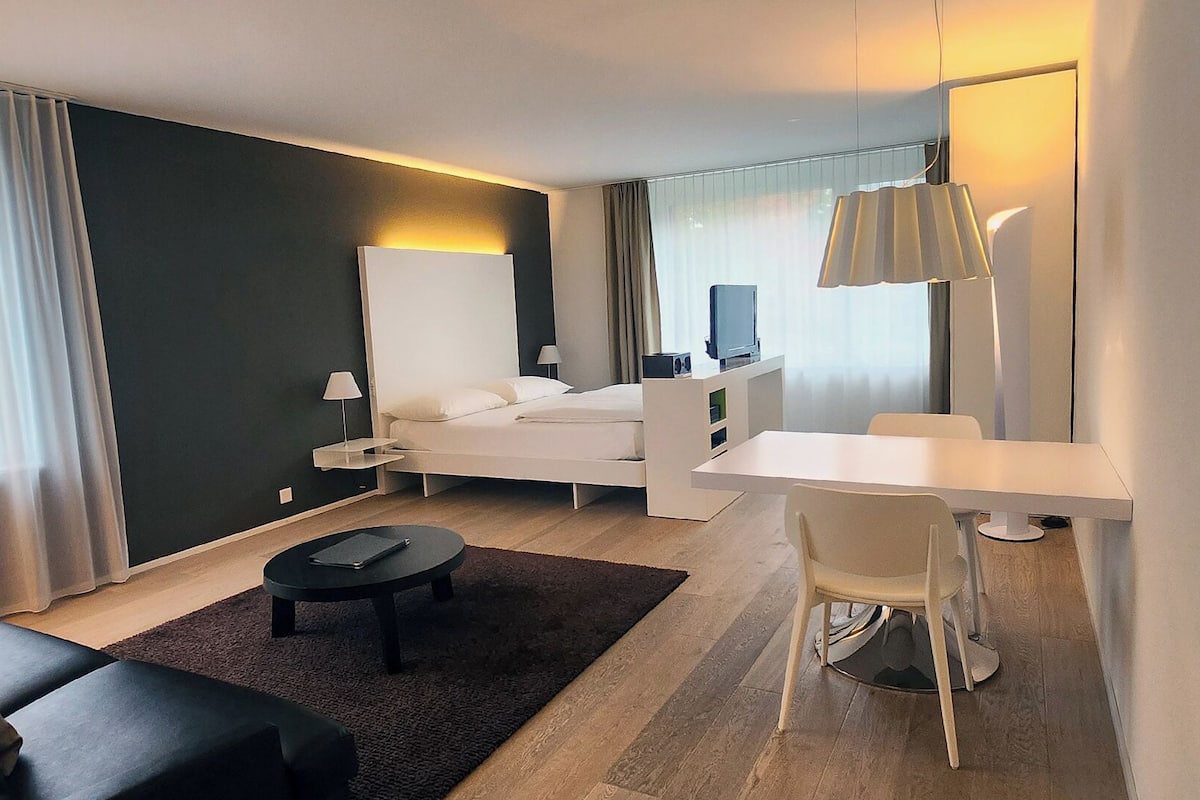 A modern hotel room with a bed, a desk, a chair, a small table, and a TV.