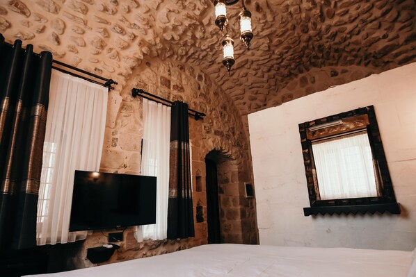 A room with a stone wall, a flat-screen TV, a bed, and a window with a curtain. - Minessa Konağı (Mardin)