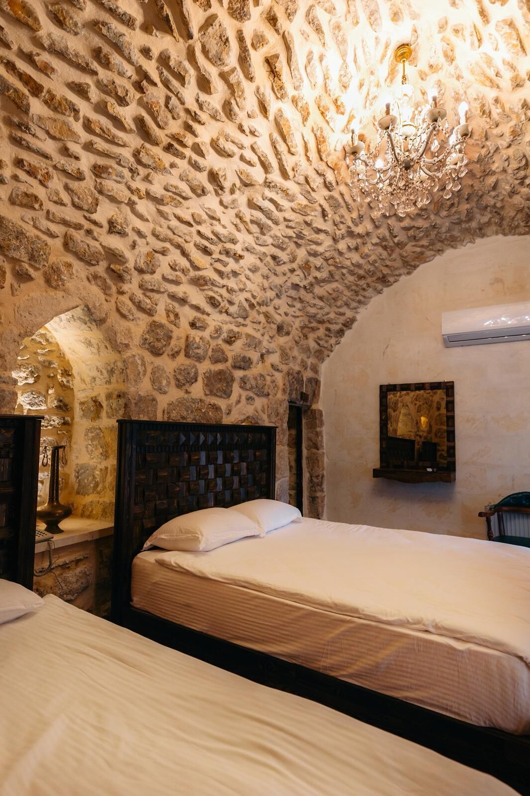 Une chambre avec un plafond en pierre, une tête de lit en bois, un lit avec une literie blanche et un climatiseur fixé au mur.