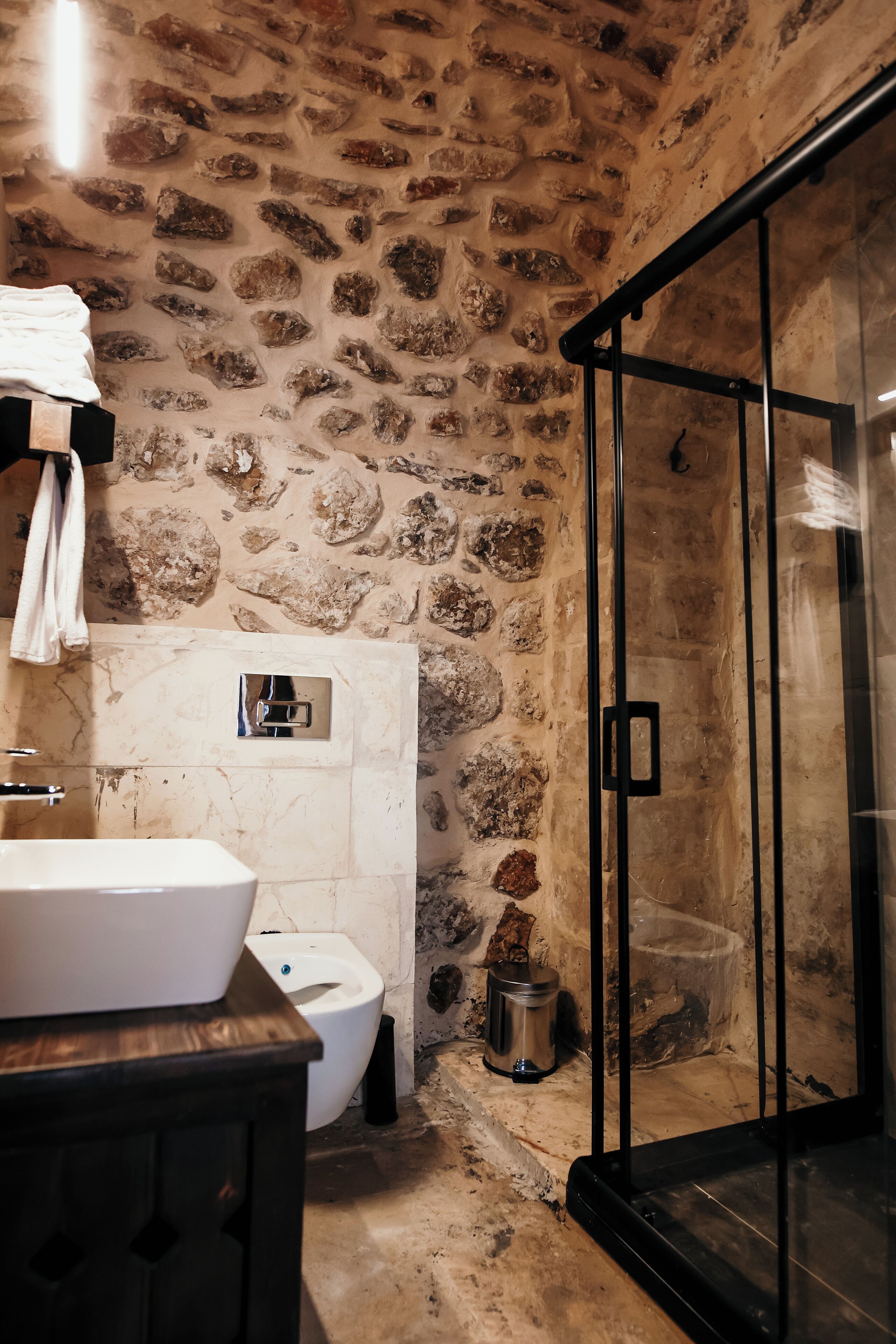 A bathroom with a stone wall, a glass shower enclosure, a white sink, and a toilet.