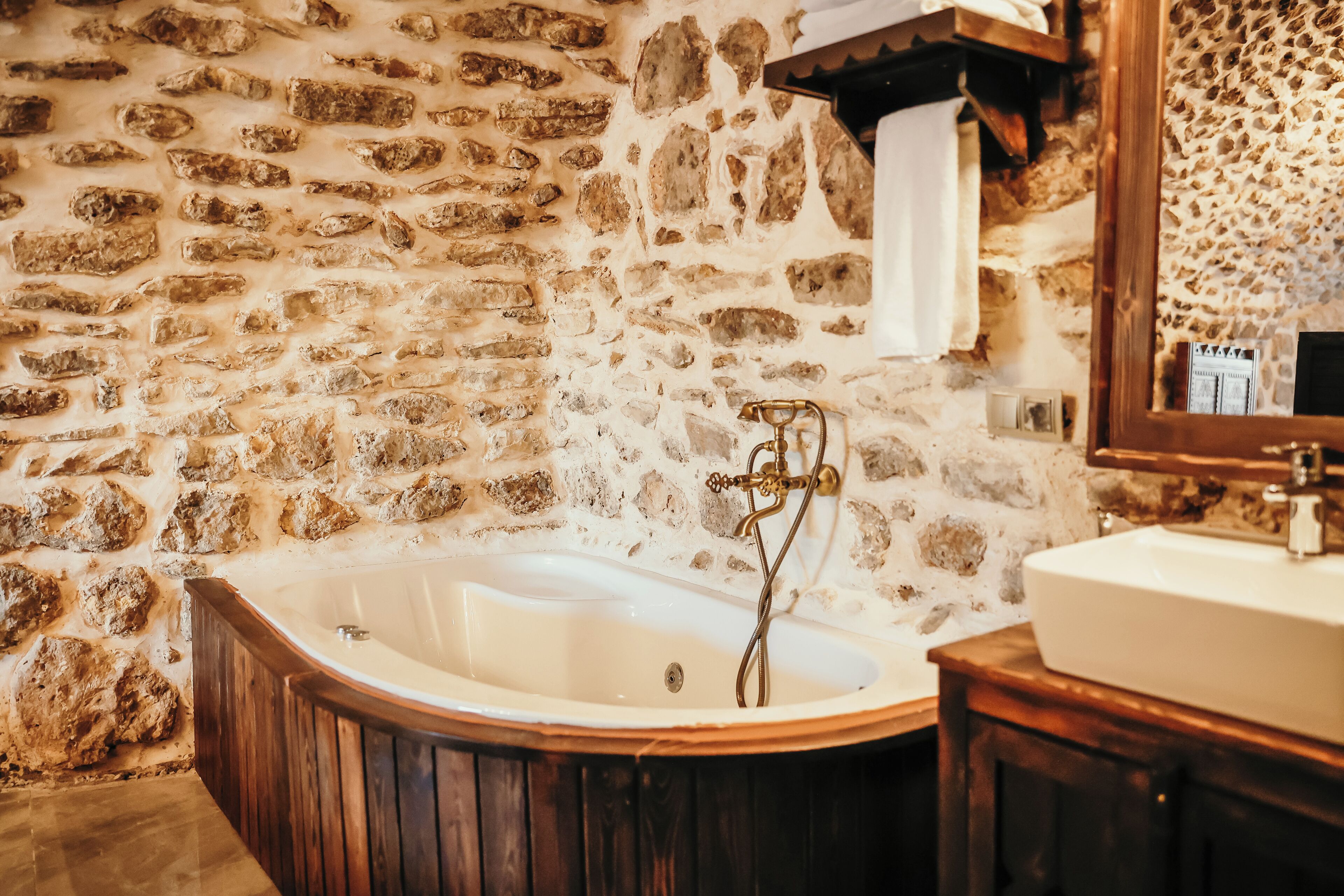 A stone-walled bathroom with a freestanding bathtub, wooden vanity, and a mirror.