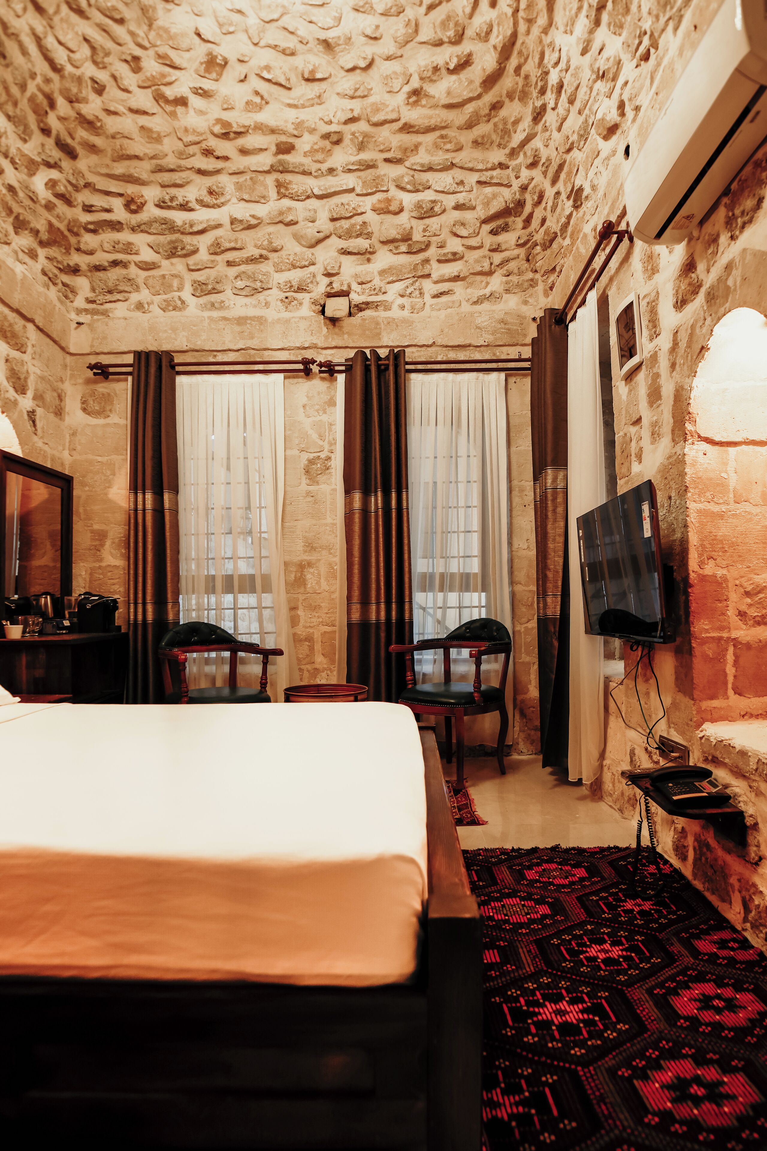 A room with a stone ceiling, a bed, a desk with a chair, a TV, and a patterned rug.