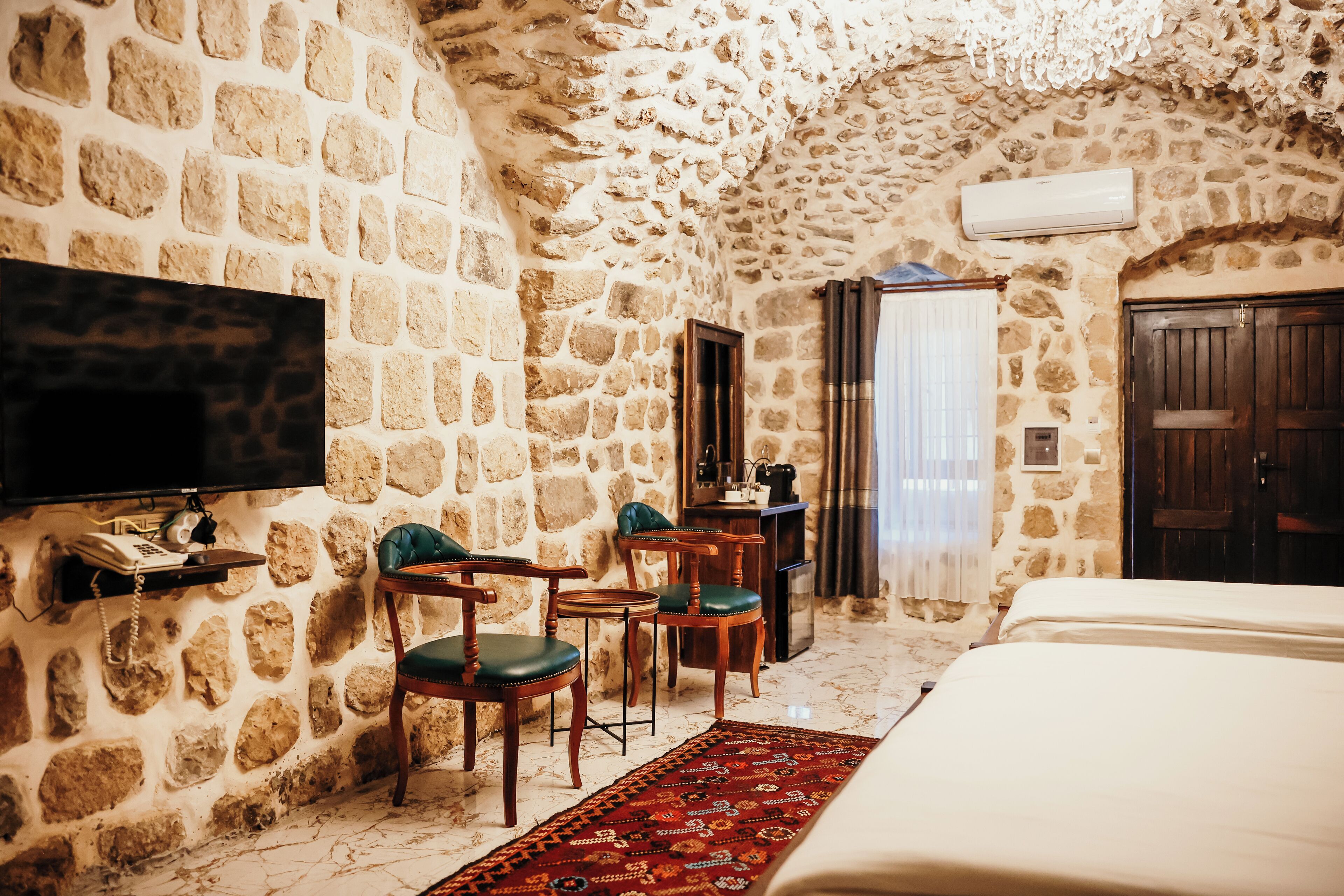 A stone-walled room with a bed, two chairs, a small table, a television, and a door.