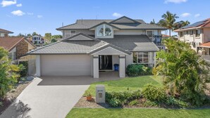 A two-story house with a grey tiled roof, a garage, and a well-maintained lawn with tropical plants.