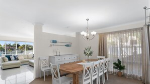 A spacious living area with a wooden dining table, white chairs, and a white sofa. There is a large window with sheer curtains, a chandelier, and a view of palm trees.