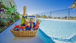 A poolside setting with a wicker basket containing snacks, a bottle of champagne, and a water bottle.