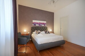 Un dormitorio moderno con un cabecero capitonado, una cama con ropa de cama blanca, un sofá gris y una pequeña mesa con una lámpara.