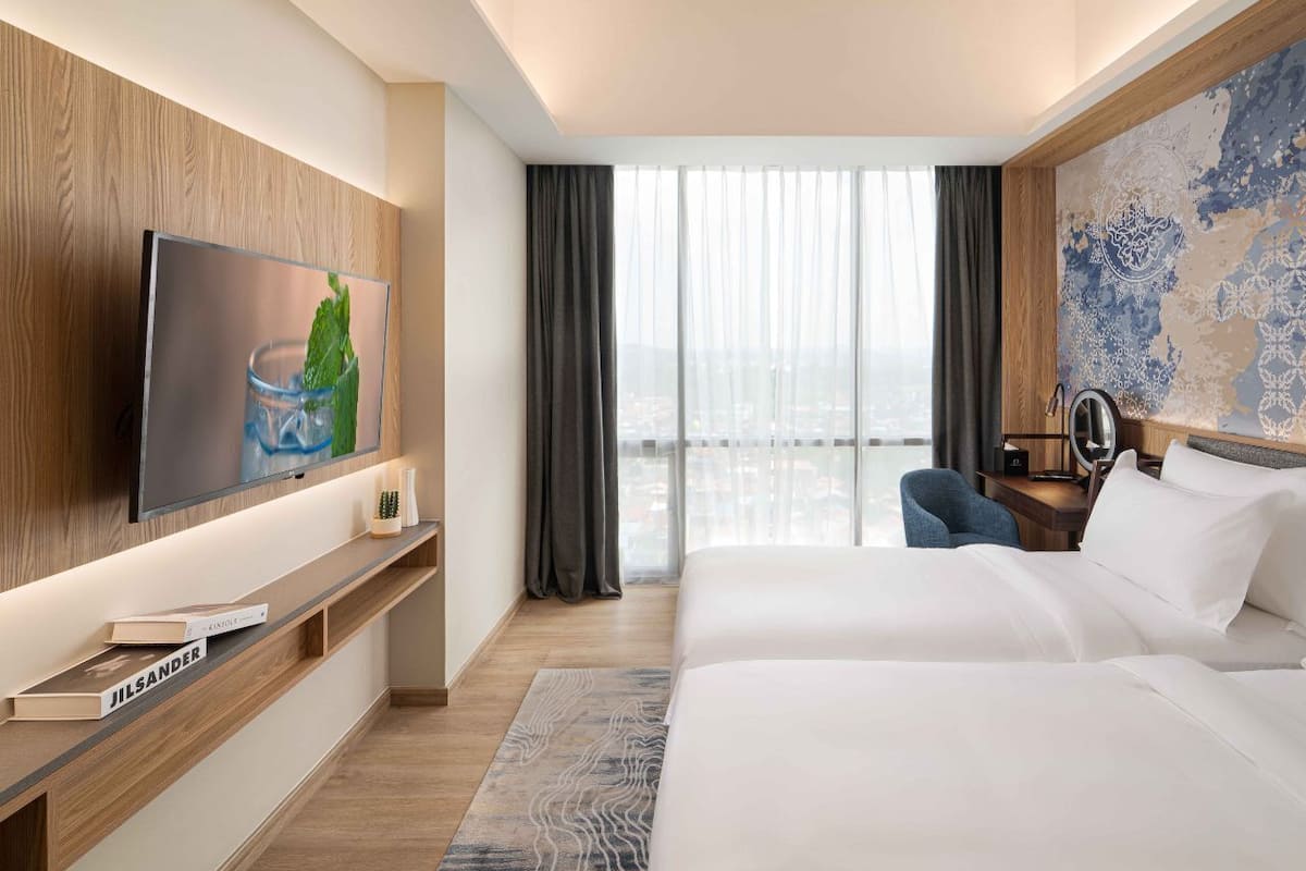 Een moderne hotelkamer met een groot bed, een flatscreen televisie, een bureau met een lamp en uitzicht op de stad door grote ramen.
