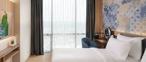 Quarto de hotel com uma cama grande, uma escrivaninha com cadeira, televisão e vista da cidade por grandes janelas.