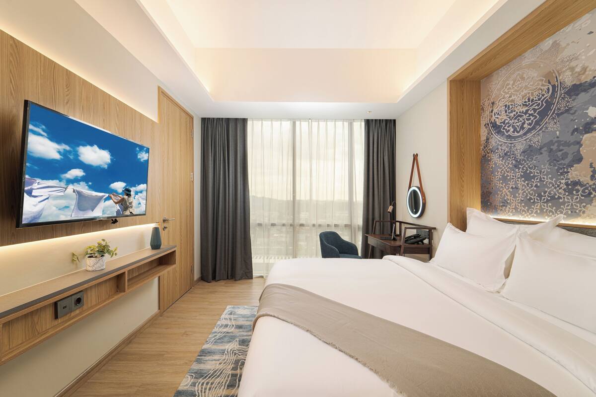 Een moderne hotelkamer met een groot bed, een flatscreen televisie aan de muur, een bureau met stoel en een groot raam met gordijnen.