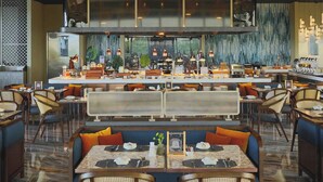 A modern restaurant with a central bar, seating with orange cushions, and a view of greenery through large windows. - Oakwood Hotel & Apartments Grand Batam (Batam)