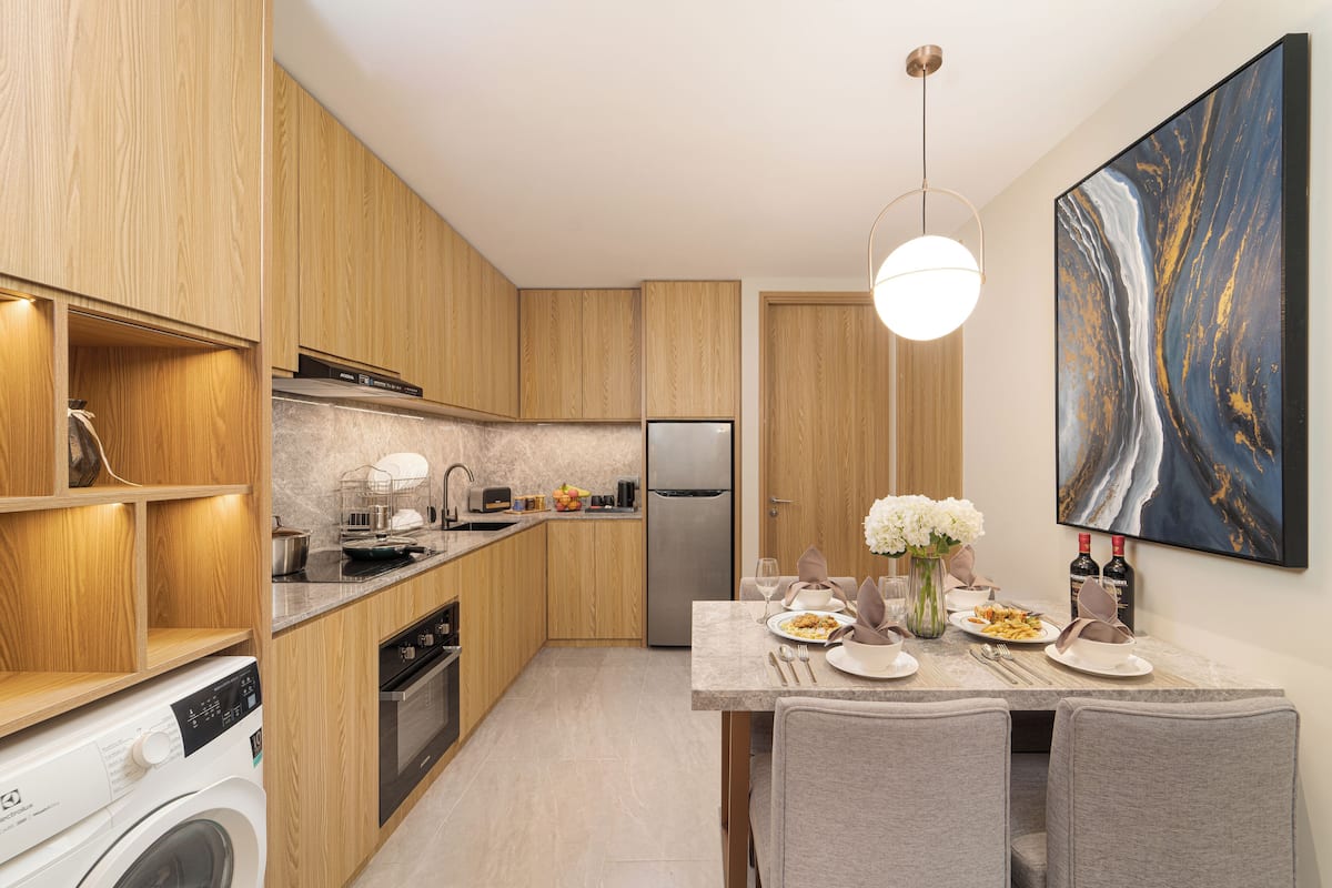 Een moderne keuken met houten kastjes, een ingebouwde wasmachine, een eethoek met een marmeren tafel en een groot abstract schilderij aan de muur.
