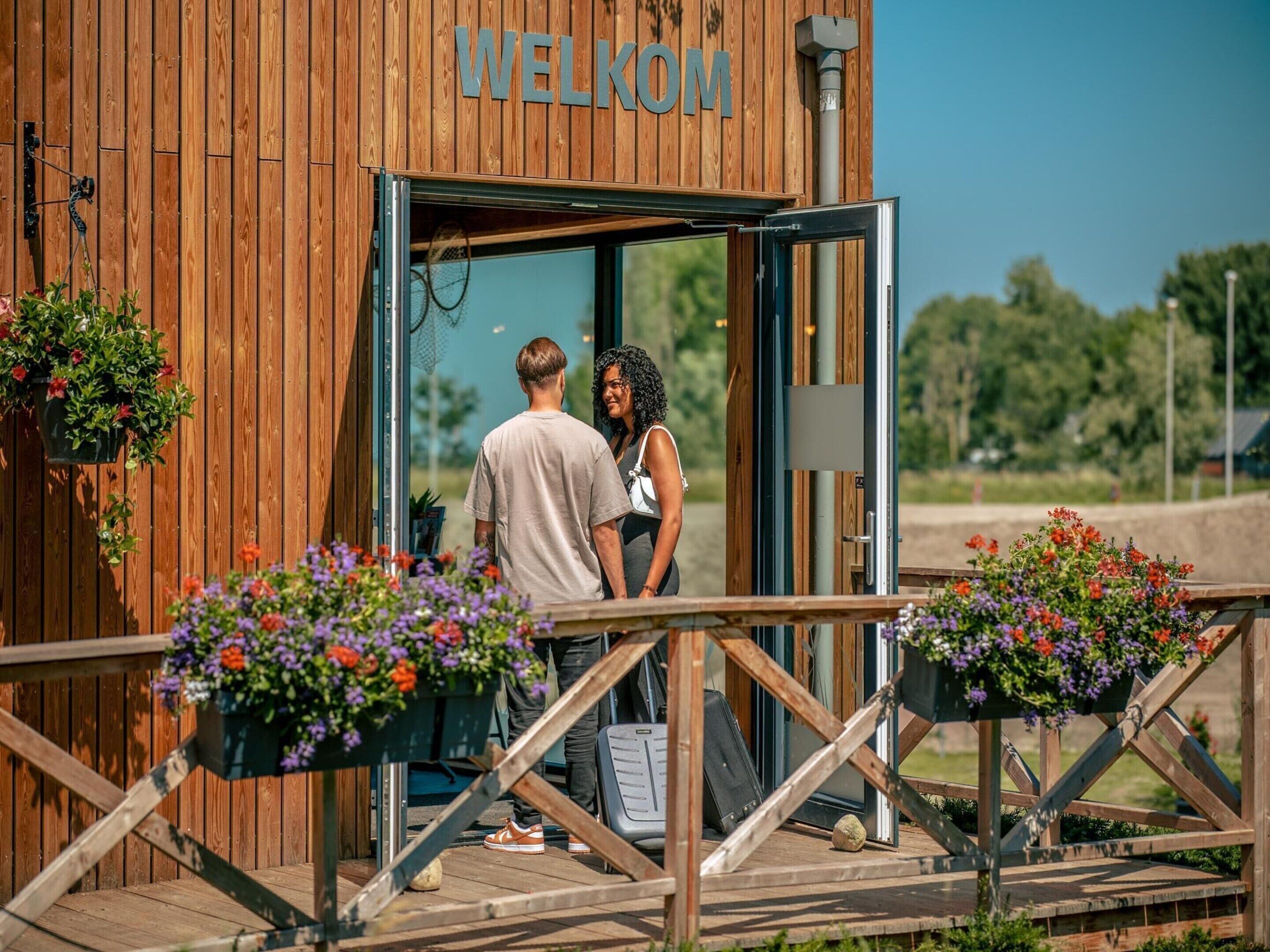 A wooden building with a 'WELCOME' sign, a wooden deck, and potted plants.