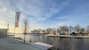 Vista sul fiume con un molo di legno, un edificio con una bandiera e una fila di case dall'altra parte dell'acqua.