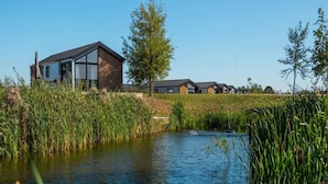 Un edificio moderno con un’ampia vetrata e rivestimento in legno, affiancato da uno specchio d'acqua, erba alta e un albero sullo sfondo.