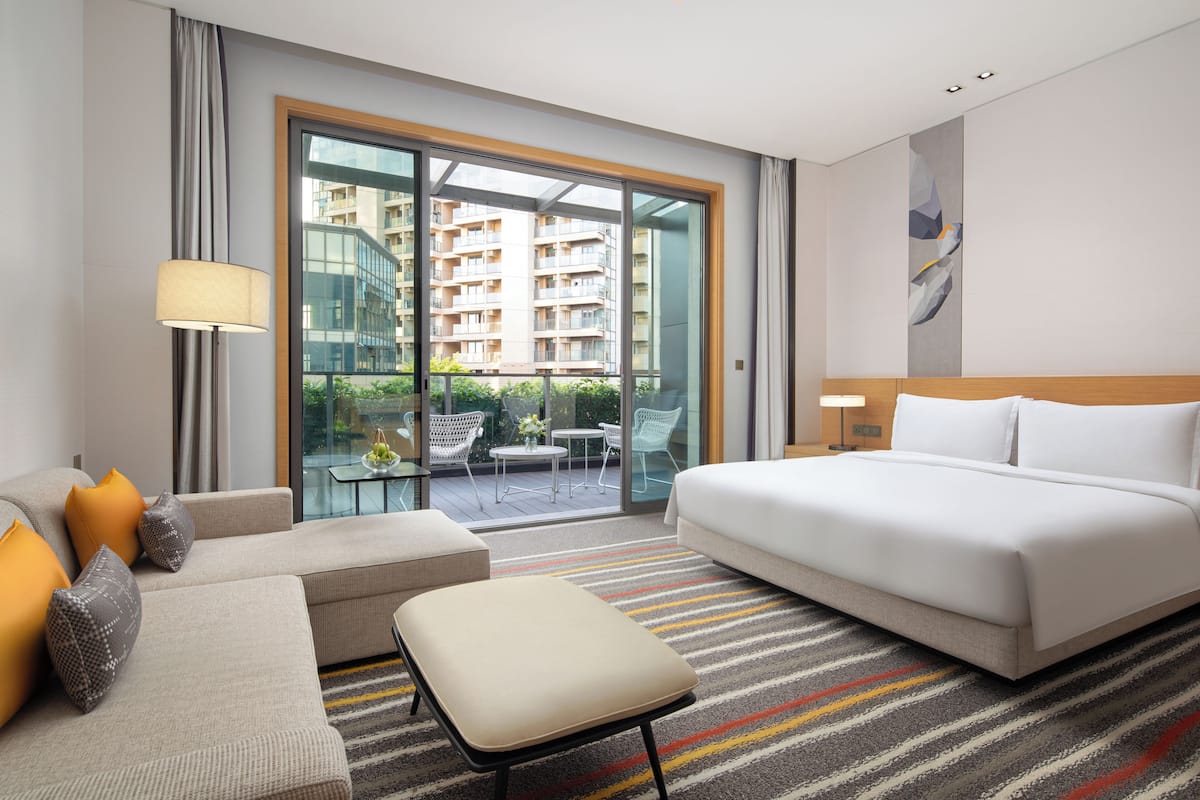 A hotel room with a large bed, a sofa, an ottoman, a glass door leading to a balcony, and a view of a building.