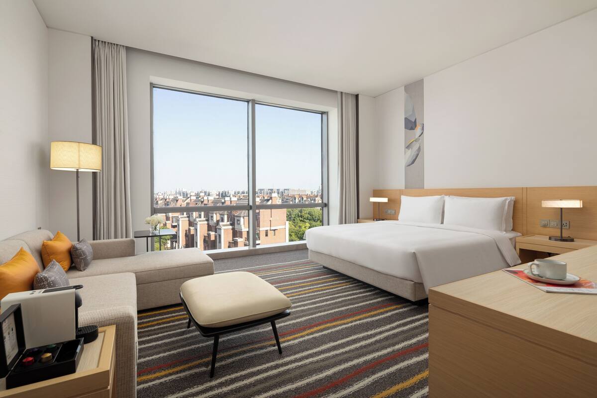 A hotel room with a large window, a bed, a sofa, a desk, and a chair.